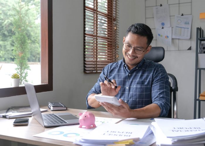 Portrait of an Asian businessman using a calculator to calculate his savings from SME operations, with a pink piggy bank as keep money concept. Portrait of an Asian businessman using a calculator to calculate his savings from SME operations, with a pink piggy bank as keep money concept.